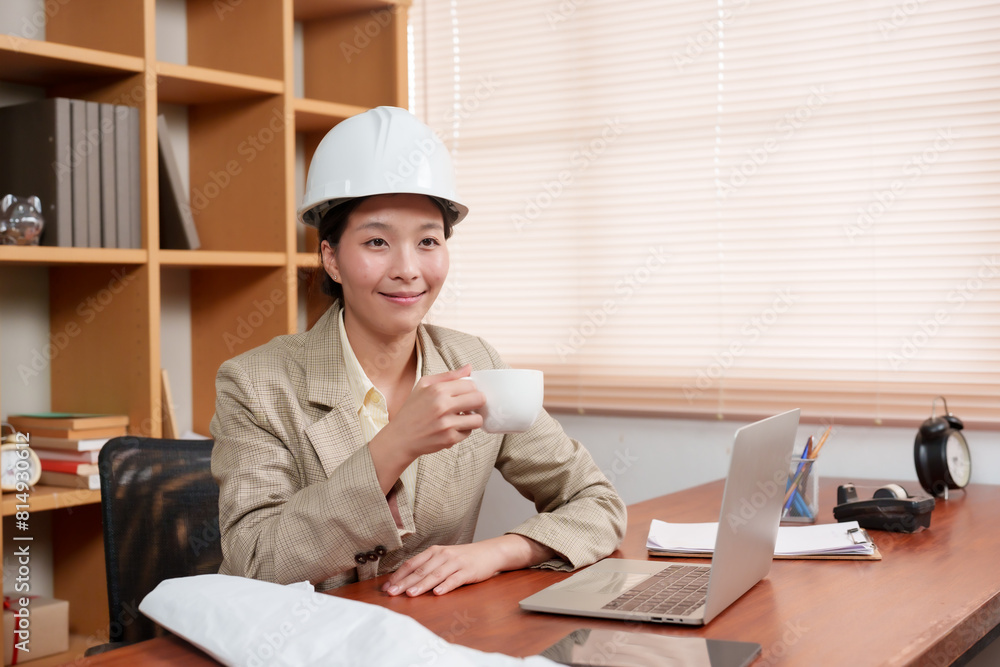 Asian female architect wearing white hard hat, sitting at desk with laptop, holding cup in modern office. Wooden shelves, green plants, documents, and clock create a professional and organized setting
