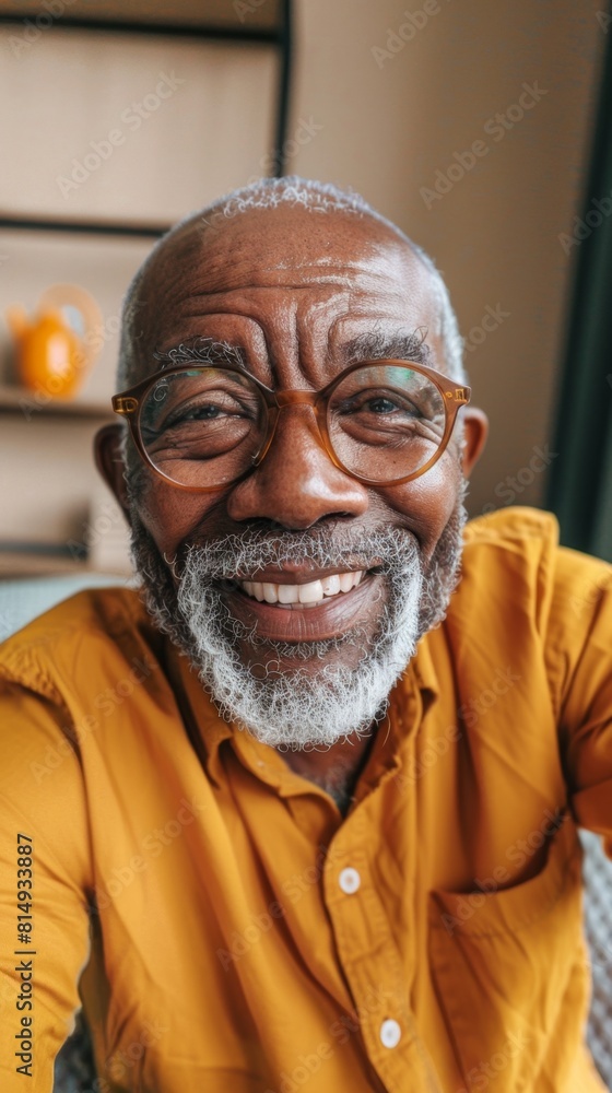Smiling elderly man with glasses and white beard wearing yellow shirt sitting in a room with a shelf in the background. Smiling elderly man with glasses and white beard wearing yellow shirt sitting in a room with a shelf in the background.