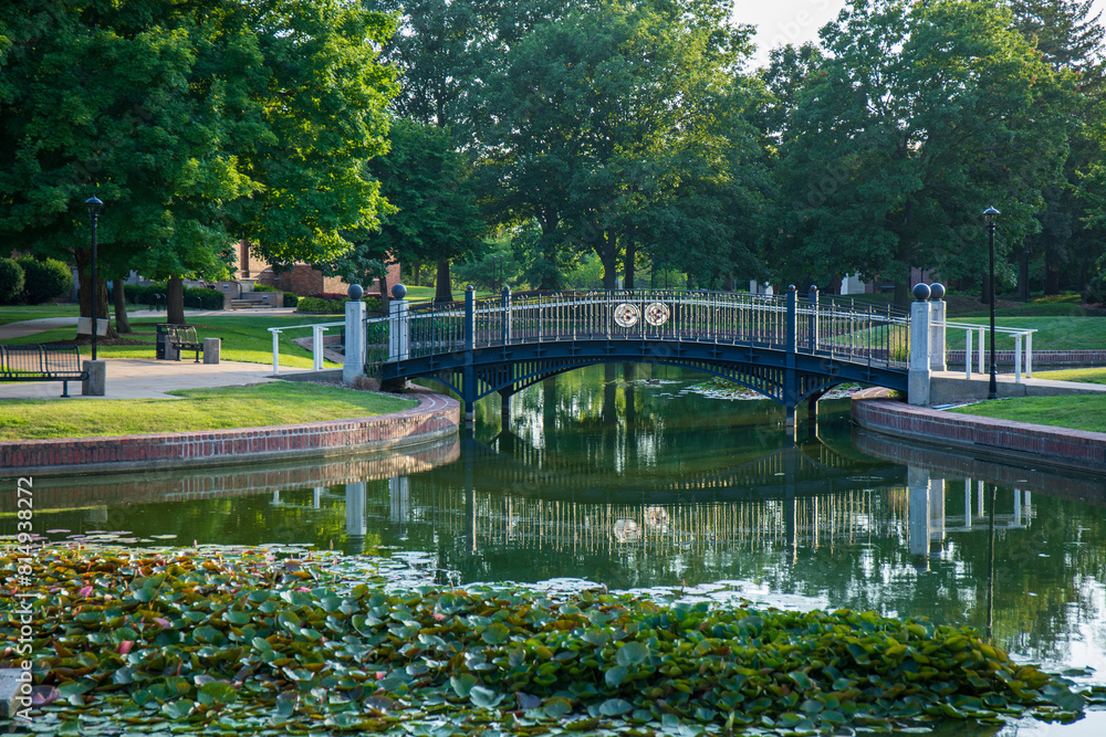 Fototapeta premium Green trees and pond with reflective surface. Pedestrian bridge over clear smooth pond. Central College campus in Pella, Iowa. 