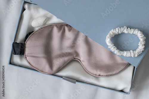 Beige sleeping mask and silk scrunchy lies in gift box on white bed.
