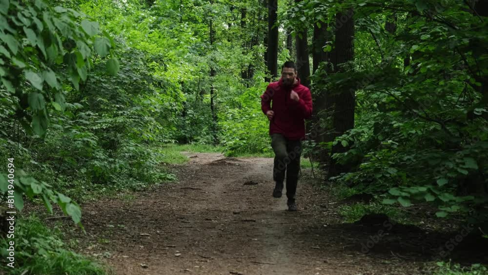 A man in a red sports jacket runs along a path in a green spring forest. 4k horizontal footage of person in motion in full height. Front view A man in a red sports jacket runs along a path in a green spring forest. 4k horizontal footage of person in motion in full height. Front view