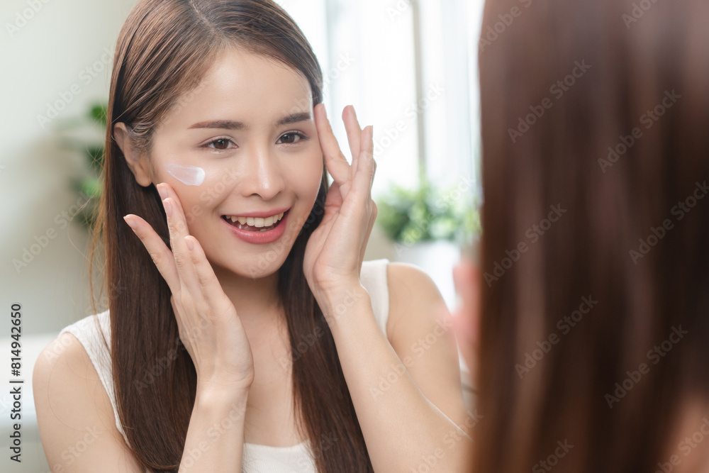 Facial beauty skin health care, smile of cute asian young woman looking at mirror, hand applying moisturizer lotion on her face, female putting cream treatment before makeup cosmetic routine at home. Facial beauty skin health care, smile of cute asian young woman looking at mirror, hand applying moisturizer lotion on her face, female putting cream treatment before makeup cosmetic routine at home.