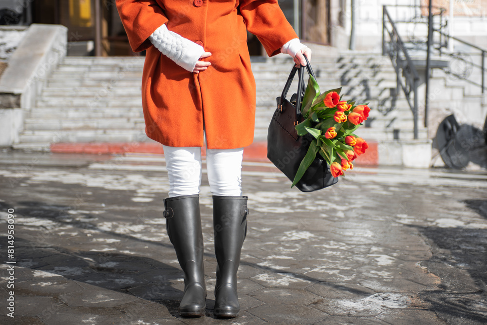 A beautiful young woman in a orange coat and rubber boots walking down the street with a bouquet of tulips in spring city A beautiful young woman in a orange coat and rubber boots walking down the street with a bouquet of tulips in spring city