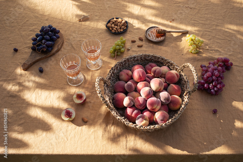 Beautiful autumn still life with a harvest of peaches in a wicker basket, two glasses of wine, grapes, honey and nuts on a table covered with a linen tablecloth. Sunny light and aesthetic shadows.