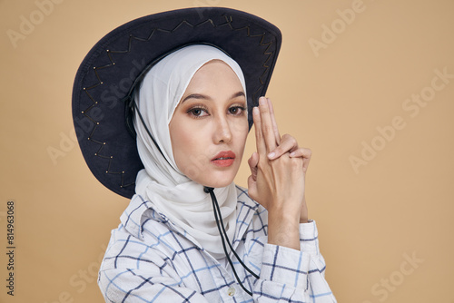 Portrait of an Asian hijab model in white shirt and blue jeans wearing cowboy hat isolated over beige background. Stylish Muslim female hijab fashion lifestyle concept. photo