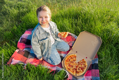 A girl in a blue denim suit sits on a blanket on green grass and holds a slice of pizza in her hand. Summer sunny day. Outdoor picnic