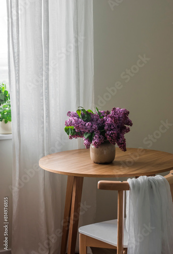 The cozy interior of the living room - a wooden table with a bouquet of lilacs in a vase  a chair by the window with linen curtains