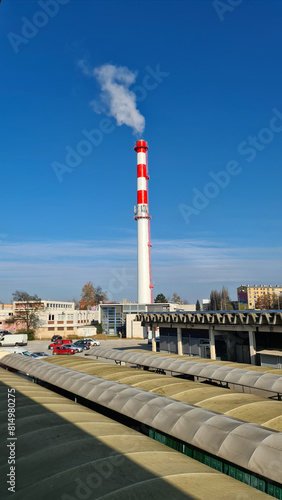 Renovated newly painted high industrial red and white chimney with multiple cell phone antennas and metal stairs overlooking small power plant building next to parking lot and old marketplace on clear