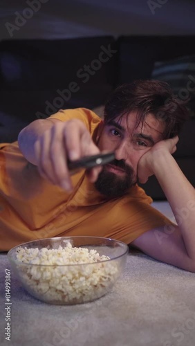 Vertical. Young European man with beard lying on living room carpet eating popcorn as he changing channel smiling happily. Millennial guy enjoying movie or series and zapping with bowl home at night photo