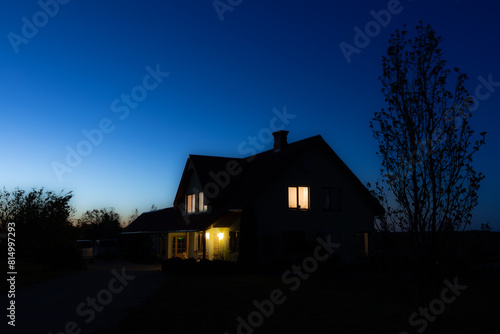 Warm lights glow from the windows of a house under the twilight sky, casting a serene ambiance over the tranquil rural setting.