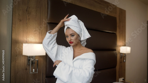 The young woman had a bath towel wrapped over her head and was wearing a bathrobe next to the bed in the hotel room. A girl in a hotel room with a towel on her head.