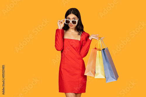 A woman wearing a vibrant red dress stands holding multiple shopping bags in her hands. bags she holds suggest she has been shopping for items, adjusting her sunglasses