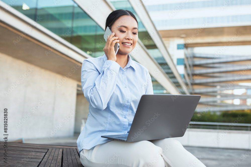 Asian woman is seated on a bench, engrossed in a phone conversation while simultaneously working on a laptop. She appears focused and multitasking efficiently in a public setting. Asian woman is seated on a bench, engrossed in a phone conversation while simultaneously working on a laptop. She appears focused and multitasking efficiently in a public setting.