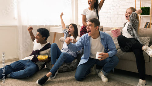 A group of friends is gathered in a cozy living room  expressing joy and excitement. Some are seated on a sofa while others sit on the floor  watching a sports game or a competitive event on TV.