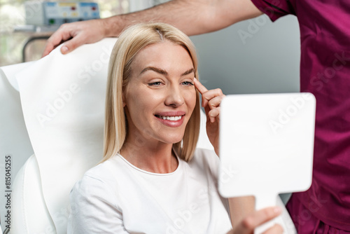 Beautiful and happy young woman sitting in medical chair and looking in the mirror. She is satisfied after successful beauty treatment with hyaluronic acid fillers or botulinum toxin injections. photo