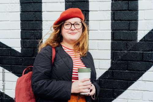 Young smiling fashion plus size woman with reusable coffee cup on painted brick background. Body positive. Female freedom. Urban street fashion style. Confident happy satisfied overweight model.