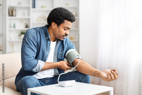 A focused young African American man sits comfortably on a couch while he wraps a blood pressure cuff around his upper arm, monitoring his blood pressure in the relaxed atmosphere of his living room. photo