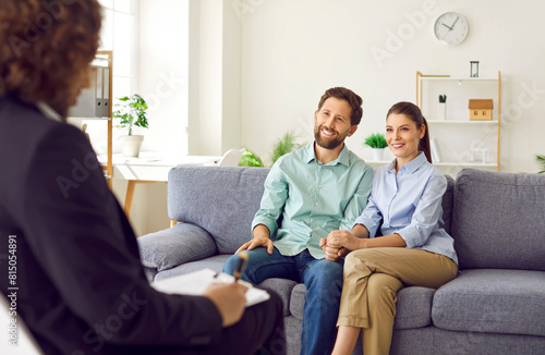Happy smiling young couple sitting on the couch at counselor's office, listening advices and smiling holding hands. Man and woman talking with male family psychologist during therapy session. photo