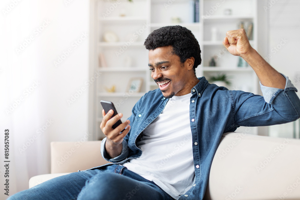 African American man is seated on a couch indoors, holding a cell phone in his hand. He appears focused on the device, with a neutral expression on his face, gesturing African American man is seated on a couch indoors, holding a cell phone in his hand. He appears focused on the device, with a neutral expression on his face, gesturing