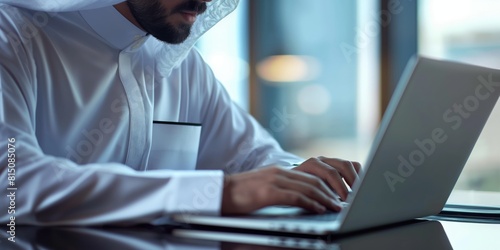 An Arab man with traditional attire focused on working with a laptop at a desk