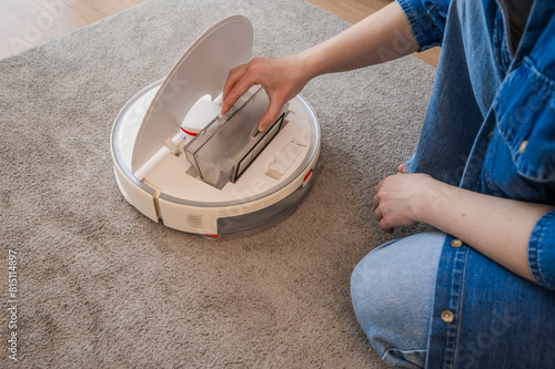 oung caucasian woman opening an automatic vacuum cleaner to clean the floor at home in living room, replace the filter. smart devices, modern technologies concept