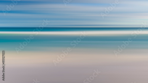 A blurry image of a beach with a blue ocean and white sand. The image has a dreamy, ethereal quality to it, as if it were a painting rather than a photograph. The water appears to be calm and still