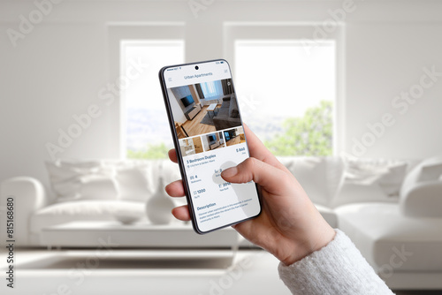 Hand holding smartphone with urban apartments search app. Living home interior in the background. Ideal for showcasing modern home search and real estate exploration on mobile devices