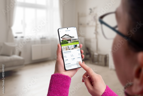 Smartphone with a residential real estate app on display in woman hands. Living room interior in the background, blending modern technology with home search