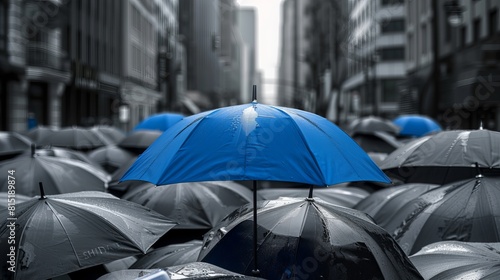 Amidst a cityscape backdrop, a blue umbrella stands out atop a cluster of gray umbrellas, symbolizing the concepts of business and safety. © Elshad Karimov