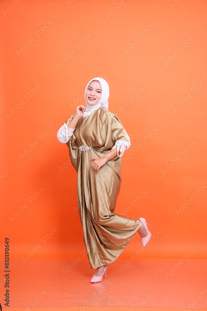 Full body of cheerful young indonesia Muslim woman wearing kaftan hijab standing attractive hands and legs up isolated orange background. Concept of Islamic religion, fashion and advertising