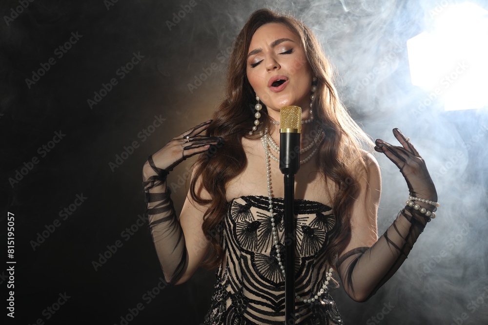 Obraz premium Beautiful young woman with microphone singing on dark background with smoke