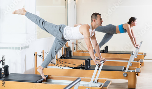 Focused young man practicing exercises on Pilates reformer to strengthen body muscles  improve posture and flexibility at group session in studio..