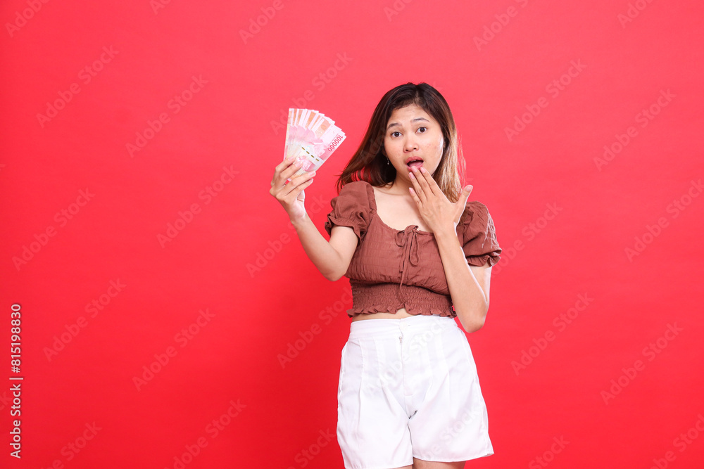 the expression of a young Asian woman shocked at the camera, covering her mouth and holding rupiah notes up, wearing a brown blouse on a red background. for transaction, technology and advertising the expression of a young Asian woman shocked at the camera, covering her mouth and holding rupiah notes up, wearing a brown blouse on a red background. for transaction, technology and advertising