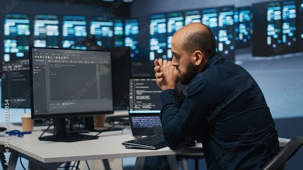 Man in server room typing code on laptop and PC, ensuring data remains shielded from threats. IT expert using notebook and computer to protect supercomputers against unauthorized access Man in server room typing code on laptop and PC, ensuring data remains shielded from threats. IT expert using notebook and computer to protect supercomputers against unauthorized access