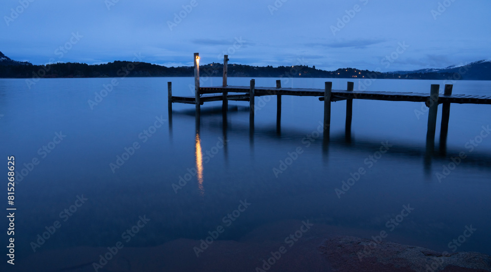 Magical view of the wooden dock in the placid lake at nightfall. Long exposure shot of the the mountains, blurry water and blue sky reflection.