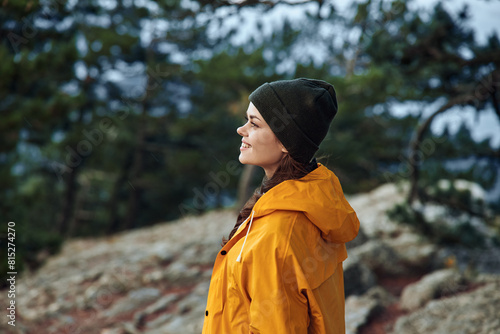 A serene young woman in a vibrant yellow raincoat standing on a rocky hill surrounded by lush trees © SHOTPRIME STUDIO