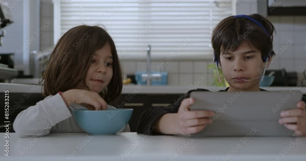 A boy with mouth braces is engrossed in his digital tablet while sitting at the kitchen table, as another child eats breakfast beside him. A boy with mouth braces is engrossed in his digital tablet while sitting at the kitchen table, as another child eats breakfast beside him.