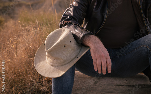 Adult man sitting on tree trunk on wild with cowboy hat on knee