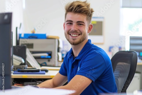 A happy man in blue polo shirt with short hair and ginger beard is sitting at his desk, working on computer inside an office space