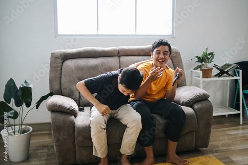 Potrait Of Funny Two Young Boys Sitting On The Sofa Laughing Out Loud While Holding Smartphone Watching Funny VIdeo at Home. 