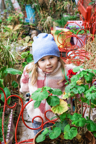 Blonde girl in a cozy jacket and blue hat standing next to a flower pot filled with red flowers. The background features lush greenery and red chairs, creating a beautiful and vibrant scene outdoors.