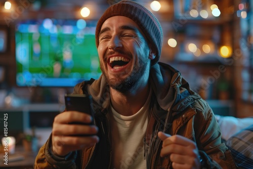 A man is happy after winning a bet. holding a mobile phone In the background there is a TV with a football match on.
