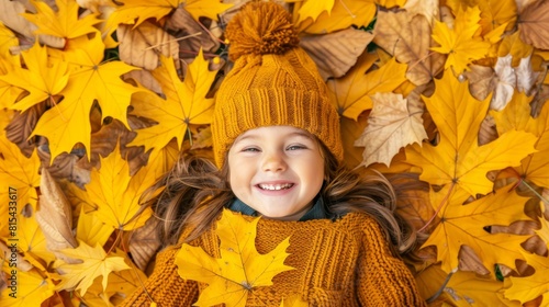  A little girl lies on a mound of leaves  dressed in a yellow knitted sweater and a hat adorned with two pom poms