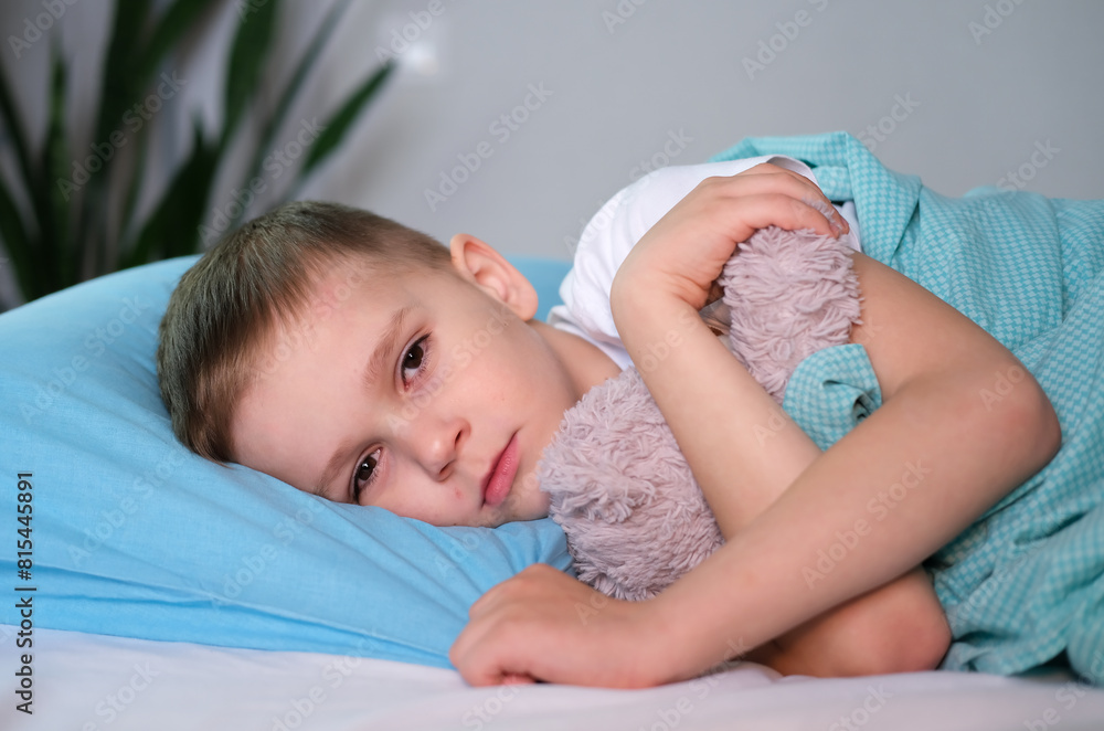 Sad little boy feels unwell, gets sick and lies in bed at home, resting in an embrace with his favorite toy. Period of flu and viral diseases. Sad little boy feels unwell, gets sick and lies in bed at home, resting in an embrace with his favorite toy. Period of flu and viral diseases.