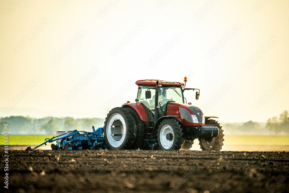 Obraz premium Farmer operates a red tractor, seeding the field against the backdrop of a setting sun