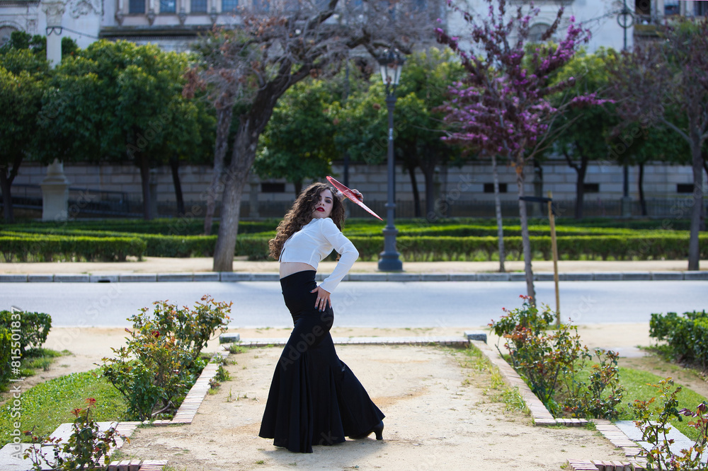 Obraz premium Beautiful woman with long curly hair, dancing flamenco with a red fan, she is in Seville, Spain. She wears long black skirt, white shirt and makes different dance poses. Flamenco, heritage of humanity
