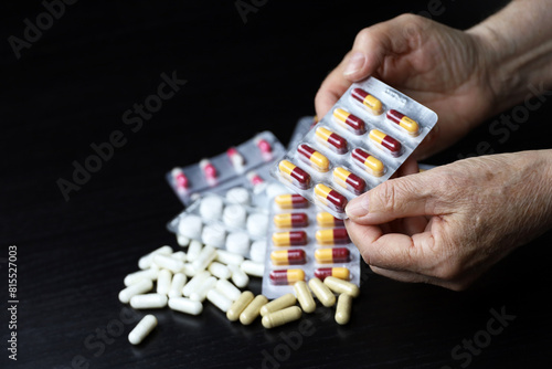 Pills in wrinkled hands of elderly woman on scattered drugs background. Medication in capsules, taking sedatives, antibiotics or vitamins in old age