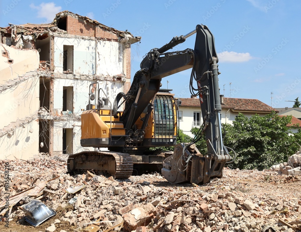 Fototapeta premium Heavy excavator with a building under demolition on behind. Front view.