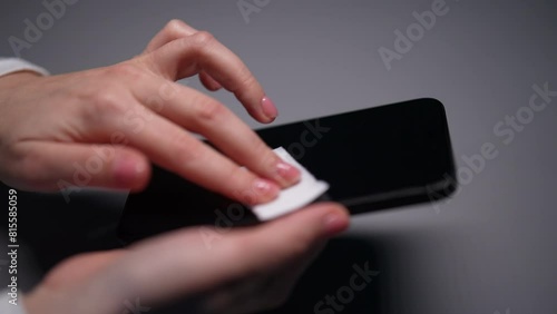 Closeup of female hand using wet wipe to clean mobile phone screen sitting at desk. Close-up of unrecognizable woman cleaning and disinfecting smartphone to kill germs in dark room, slow motion.