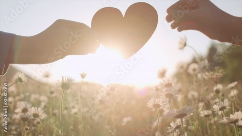 Close up of mother and child daughter hands holding small red paper heart near flowers on background beautiful sunset or sunrise at warm summer day. Concept of symbol love, parenthood and happy family